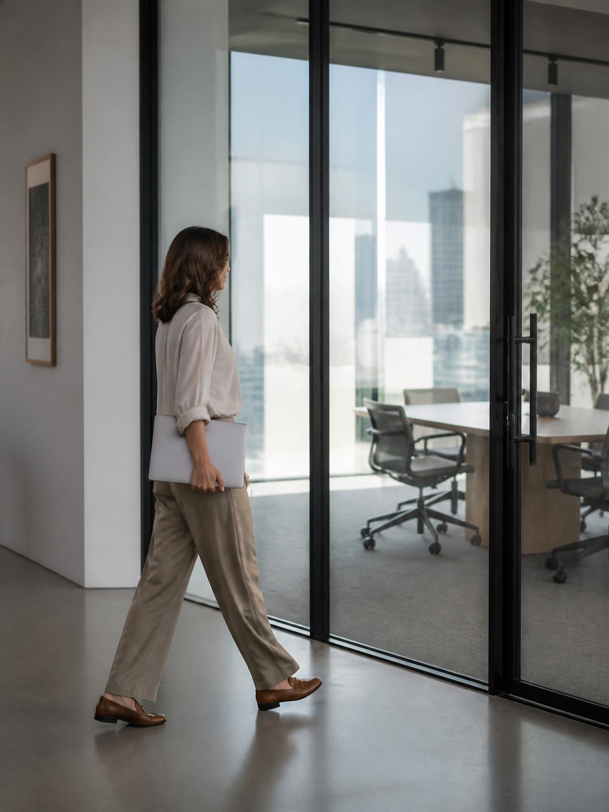 A woman carrying a laptop walks past a glass meeting room with a city skyline visible beyond