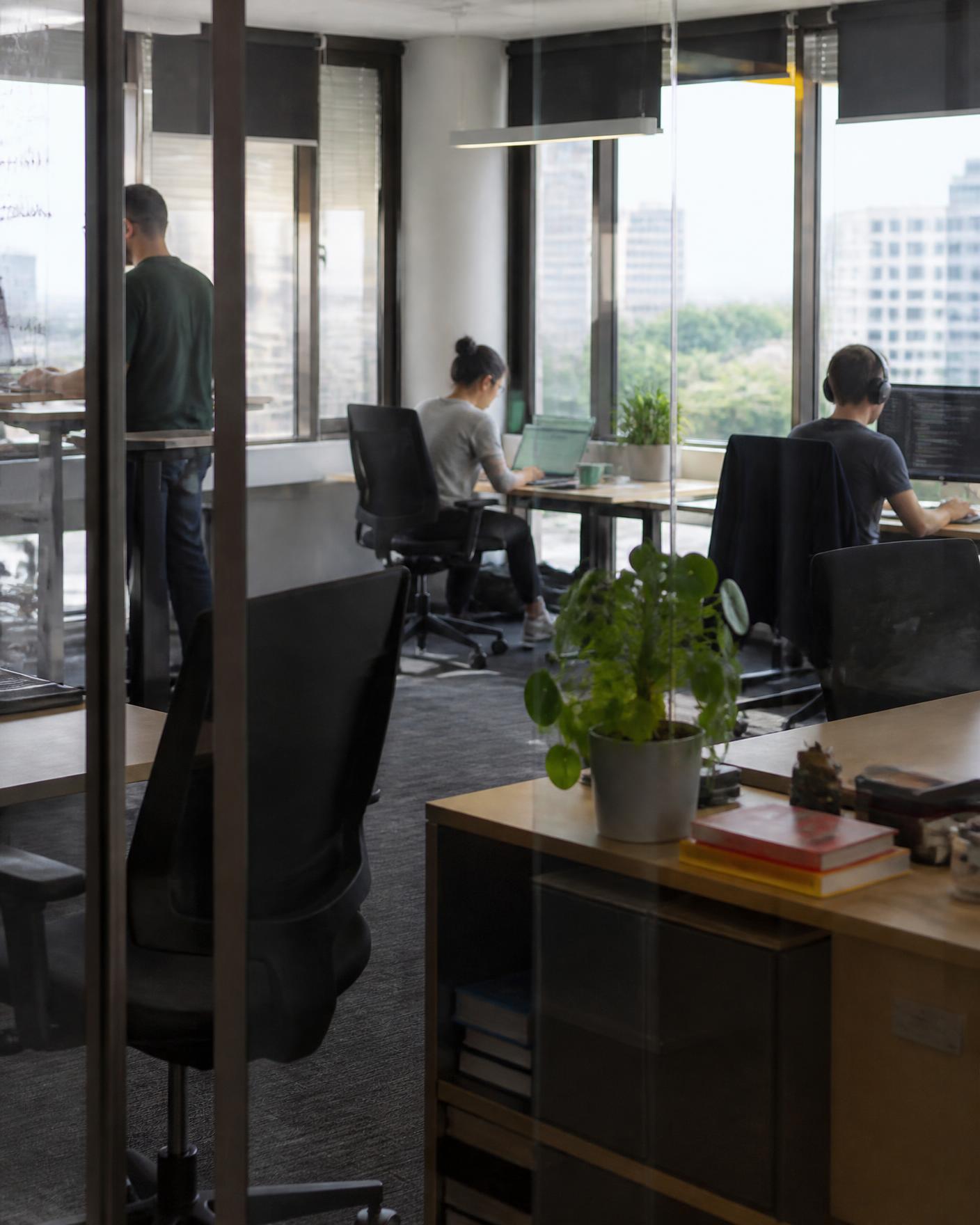 Three people working independently in a glass-walled office overlooking the city, with laptops, monitors and plants on desks