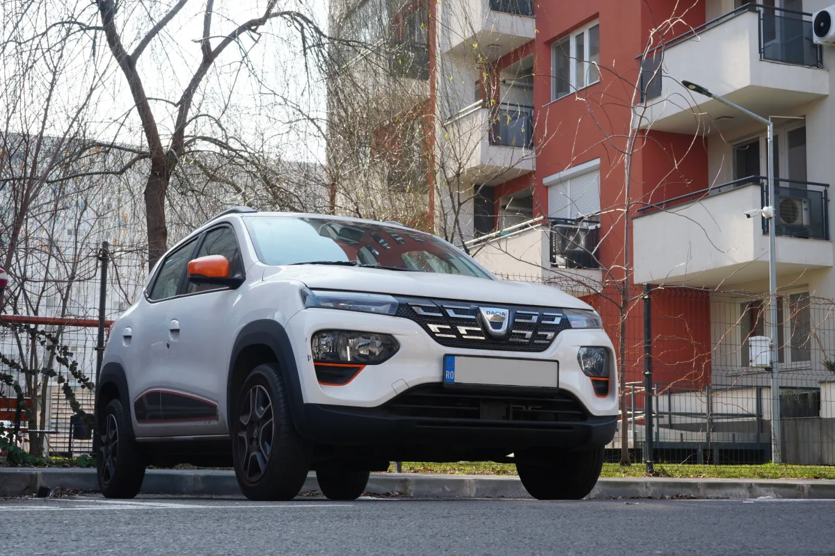 A white Dacia Spring, an all-electric city car, is parked on a street in front of apartment buildings.
