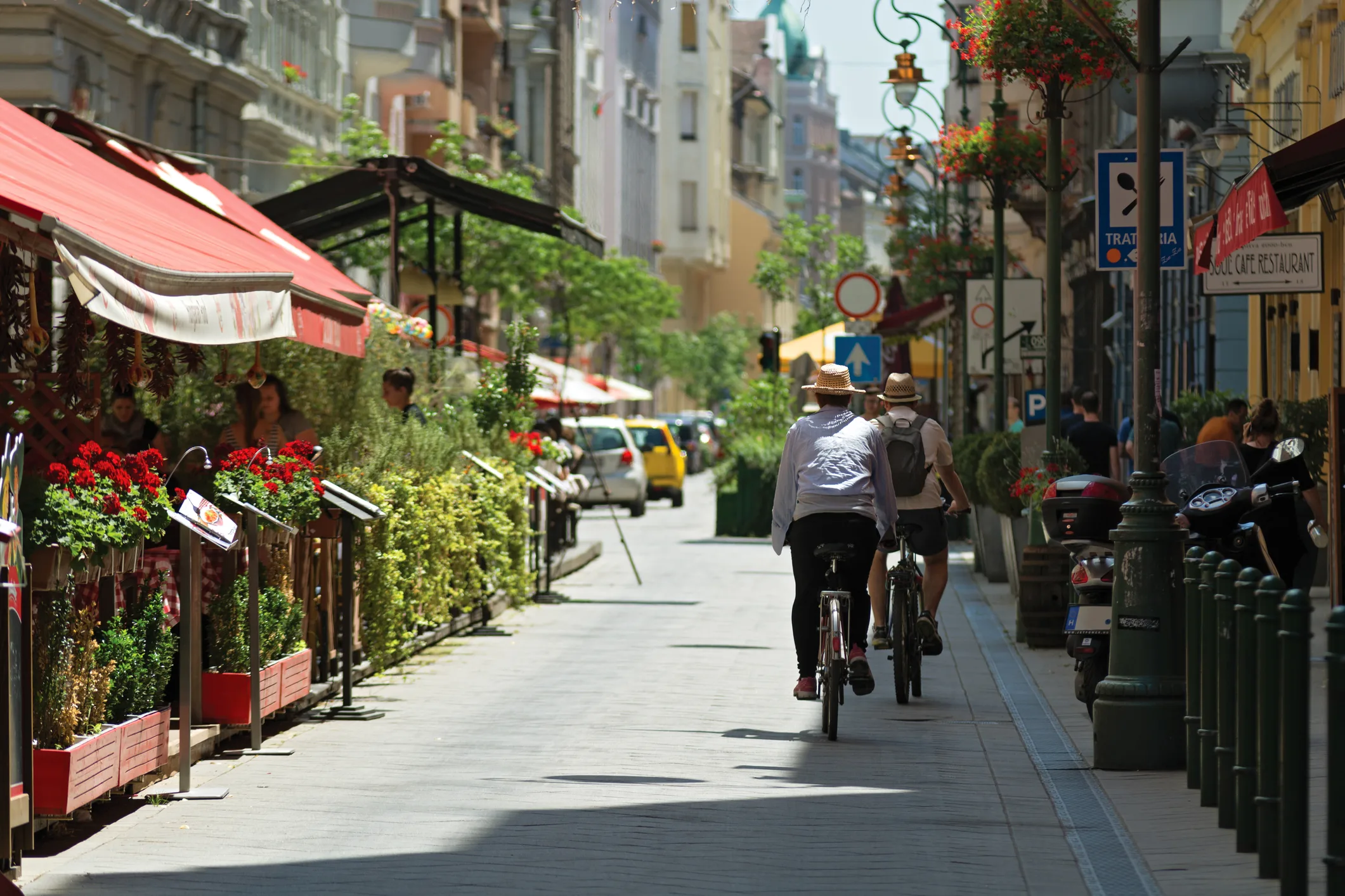 Tourists cycling through a lively pedestrian street in Budapest, Hungary, surrounded by outdoor cafés, red awnings, and summer greenery