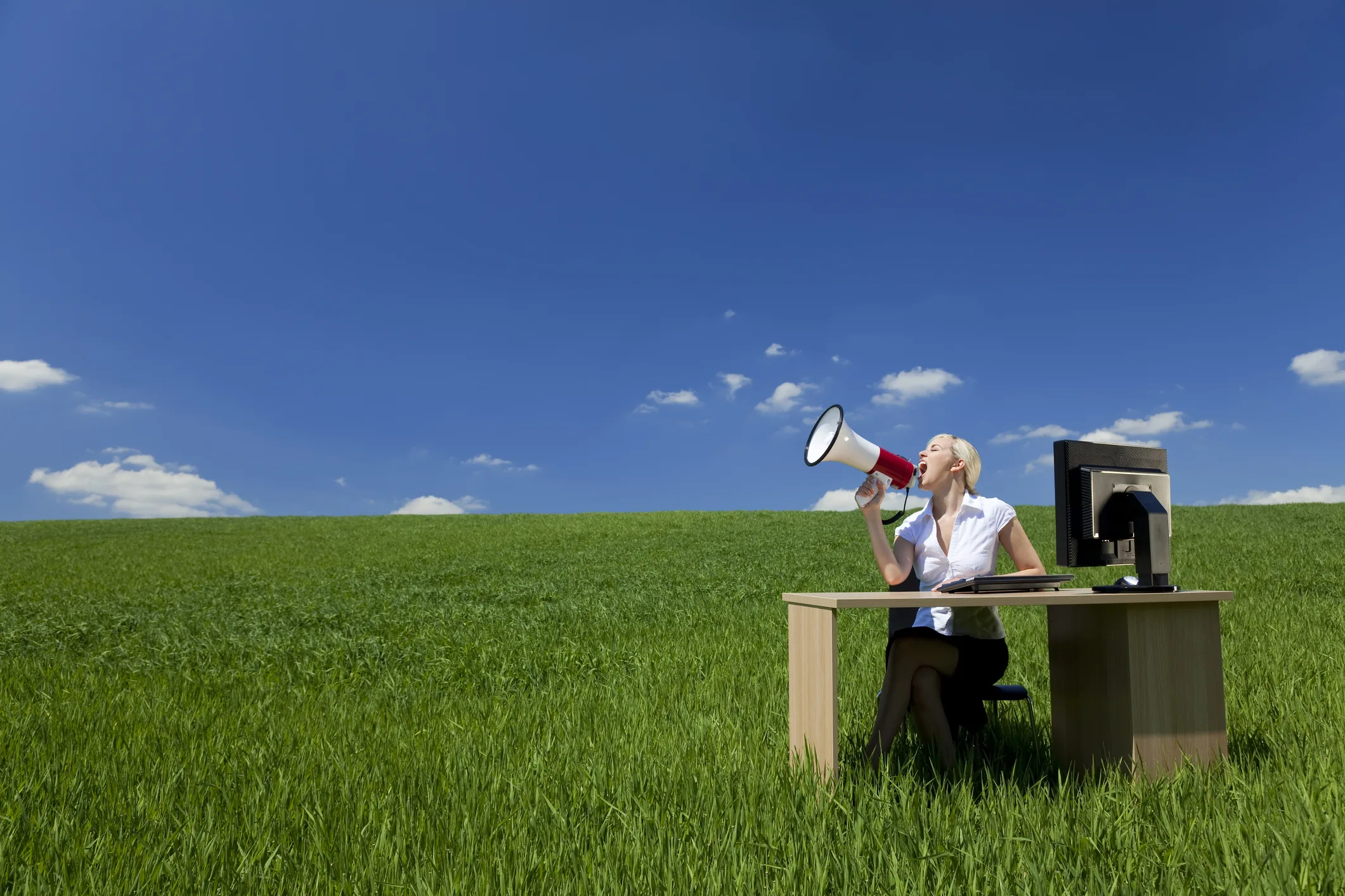 Businesswoman sitting at a desk in a grassy field using a megaphone, symbolizing modern corporate communication and the evolving role of press releases in a digital media landscape