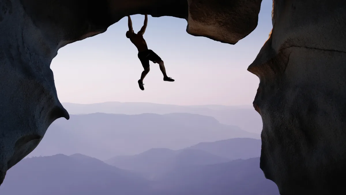 Man hanging from a cliff edge, symbolizing courage, risk-taking, and the journey of personal reinvention.