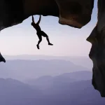 Man hanging from a cliff edge, symbolizing courage, risk-taking, and the journey of personal reinvention.