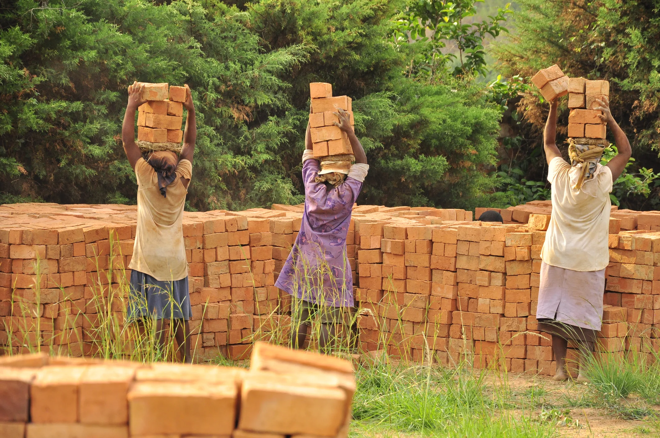 Women carrying stacks of bricks at a construction site, highlighting gender inequality in manual labor and the debate over ‘real jobs’ versus so-called ‘non-jobs’ in modern economies.